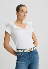 Tricou alb cu mânecă scurtă, cu decolteu volănat, asortat cu blugi din denim albastri și o curea albă cu model, care are cataramă aurie.