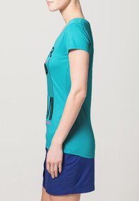 Camiseta de algodón en color verde azulado con mangas cortas, que presenta un diseño gráfico en negro y rosa en la parte delantera, combinada con una falda azul.