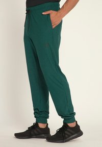 Groene sportieve joggers met een elastische tailleband, een trekkoord, zijzakken en een subtiele logo-ontwerp, gecombineerd met zwarte sportschoenen.