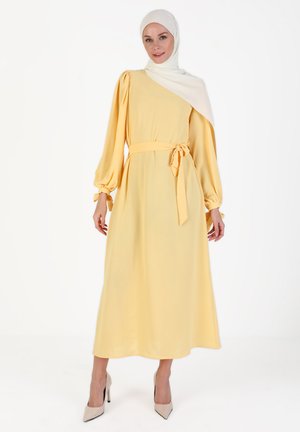 Yellow long-sleeve dress with a belted waist, asymmetric design, soft fabric, and balloon sleeves. Paired with a white hijab and heels.