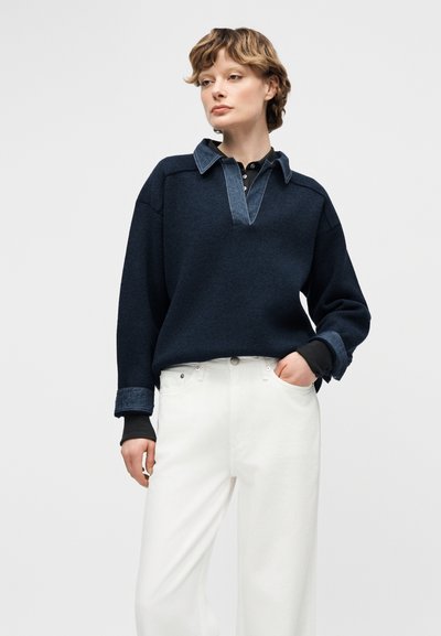 Personne portant un pull bleu marine avec un col et des poignets en denim par-dessus un haut noir, combiné avec un pantalon blanc taille haute, debout les mains dans les poches.
