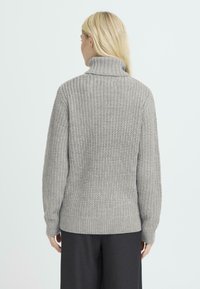 Grijze, chunky gebreide trui met een hoge kraag en textuurstof. Heeft ribgebreide boorden en onderrand, een losse pasvorm en wordt gedragen met zwarte broeken.