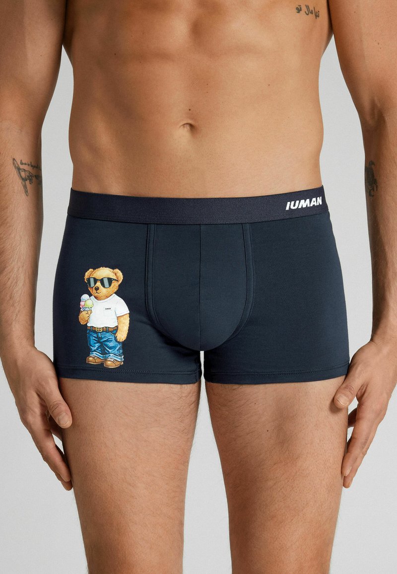 Männlicher Torso, der schwarze Boxershorts trägt, mit einem Cartoon-Bären in Sonnenbrille, der auf dem linken Oberschenkel ein Eis hält.