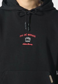 Sudadera negra con texto bordado en rojo "PAS DÉ MONACO 1988" y "Atelier Racing". Cuenta con cordones y un bolsillo frontal. Hecha de material suave.