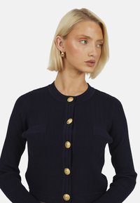 Marineblauwe geribde cardigan met een ronde halslijn, gouden knopen en twee kleine borstzakken. De stof is gestructureerd en aansluitend.