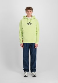 Sudadera verde claro con cordones y bolsillo frontal, con el logo de "Alpha Industries". Combinada con jeans azul oscuro y zapatillas deportivas.