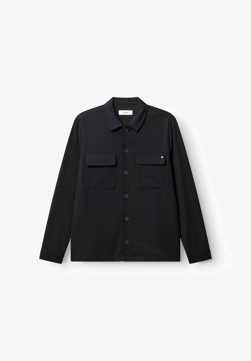 Chemise noire à manches longues en guise de veste, col pointu, boutonnée à l'avant, avec deux poches à rabat sur la poitrine, étalée à plat sur un fond blanc.