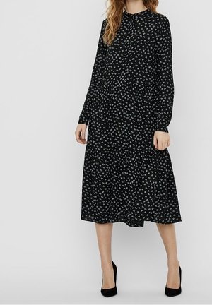 Kvinde iført en sort midi-kjole med lange ærmer med små hvide blomsterprint og sorte højhælede sko mod en ensfarvet baggrund.