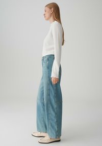 Weißes, strukturiertes Langarmoberteil, hochtaillierte hellblaue weite Jeans und beige Loafers mit schwarzen Sohlen. Schlichtes, minimalistisches Design.