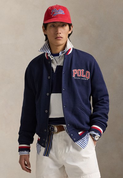 Homme portant une casquette rouge Polo, une veste Polo Ralph Lauren bleu marine, une chemise rayée, un short blanc et une montre, main dans la poche.