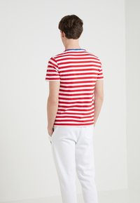 Červené a bílé pruhované tričko s modrým výstřihem, přiléhavý střih, krátké rukávy; nošeno s bílými kalhotami, zobrazeno zezadu.