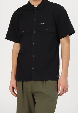 Zwart kortärmelig overhemd met kraag, twee borstzakken, drukknoopsluiting en een klein logolabel. Draagt het met olijfgroene broek.