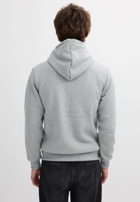 Sudadera gris con capucha ajustable, puños y dobladillo de canalé, hecha de una tela suave; llevada con jeans oscuros, vista desde la parte de atrás.