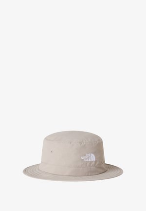 Sombrero tipo pescador de tela color beige claro, con ala ancha, detalles de costuras y un logo blanco en la parte frontal.