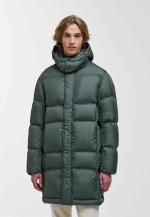 Junge Person mit mittellangen Haaren, die einen langen, dunkelgrünen, gesteppte Wintermantel mit hohem Kragen und Kapuze vor einfarbigem Hintergrund trägt.