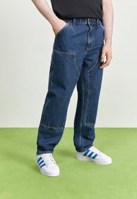 Jeans aus Denim mit lockerer Passform, dunkelblauer Farbe, Fronttaschen und kontrastierenden gelben Nähten. Kombiniert mit weißen Sneakers mit blauen Streifen.