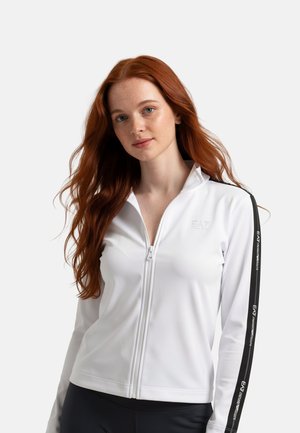 Giovane donna con lunghi capelli rossi che indossa una giacca bianca con zip, con strisce nere e testo sulle maniche.