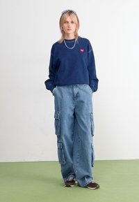 Marinegenser med en liten rød hjerte-logo, paret med lyseblå cargo-bukser med flere lommer og en avslappet passform. Brune joggesko.