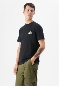 Un jeune homme portant un t-shirt noir avec un logo blanc se tient devant un fond blanc, vêtu de pantalons cargo vert olive et affichant un tatouage floral.