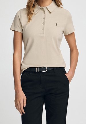 Vrouw draagt een beige korte mouw polo met knoopsluiting en zwarte broek met zwarte riem, hand in zak.