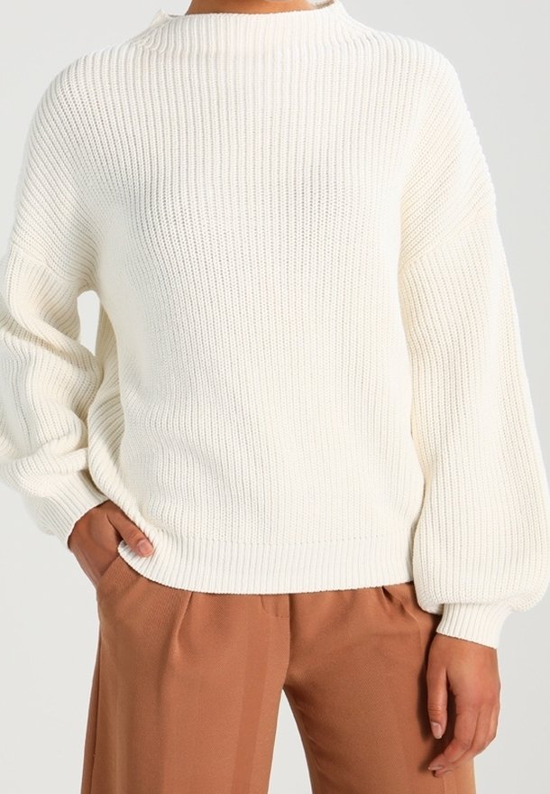 Weißer, gestrickter Pullover mit geripptem Muster, hohem Kragen und Ballonärmeln, kombiniert mit braunen Hosen.