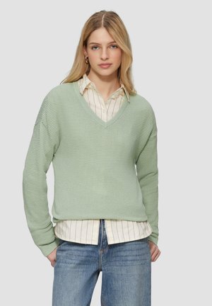 Pull en maille vert clair avec un col en V, des manches longues et une coupe ample, superposé sur une chemise blanche à rayures à boutons et un jean bleu.
