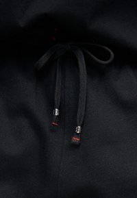 Tissu noir avec un cordon noir noué doté d'embouts métalliques argentés et de petites coutures rouges près des extrémités.