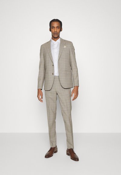 Costum cu carouri de culoare maro deschis, cu o croială slim, care include un sacou cu un singur rând de nasturi și pantaloni asortați. Însoțit de o cămașă albă și pantofi maro.