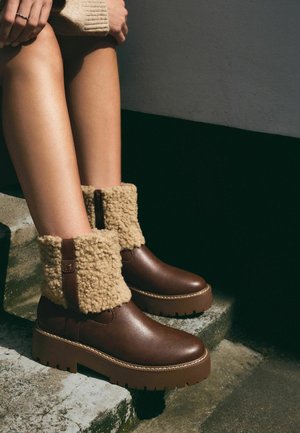 Bottes chevilles en cuir marron avec un bord en fourrure beige duveteux, semelle épaisse et détails cousus le long du bord, placées sur une surface en pierre.