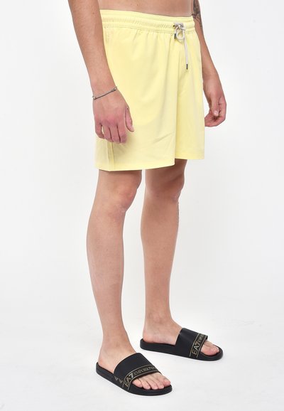 Pantaloni da bagno gialli con vita elastica e lacci. Abbinati a infradito nere con logo del marchio in oro. Design semplice.
