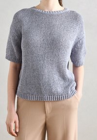 Heller grauer kurzärmeliger Strickpullover mit Rundhalsausschnitt, geripptem Saum und lockerer Passform, kombiniert mit tanfarbener Hose. Strukturiert gewebter Stoff sichtbar.