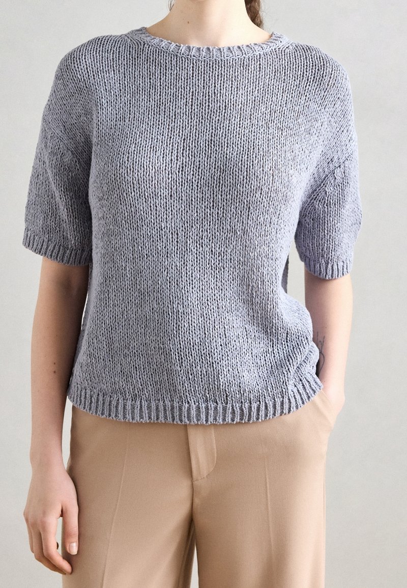 Heller grauer kurzärmeliger Strickpullover mit Rundhalsausschnitt, geripptem Saum und lockerer Passform, kombiniert mit tanfarbener Hose. Strukturiert gewebter Stoff sichtbar.