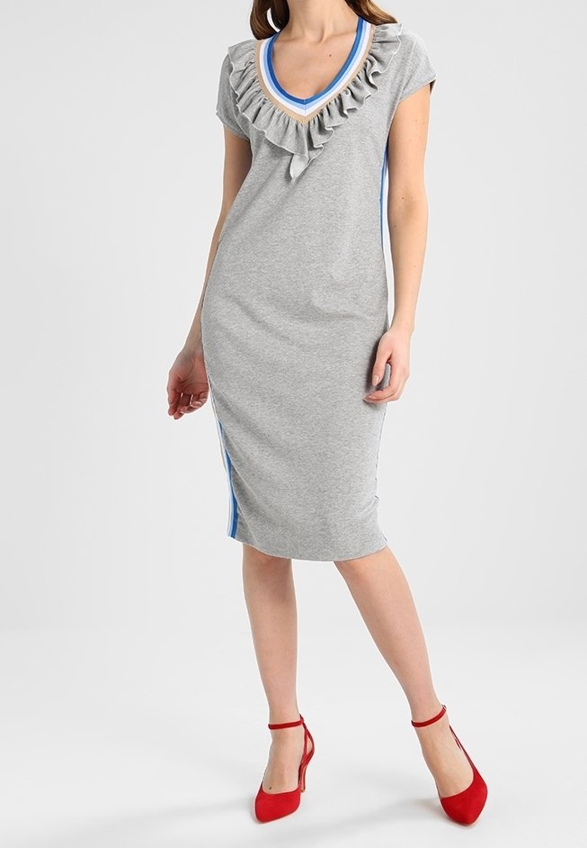 Kvinna i en grå knälång klänning med volangad V-halsringning och blåa ränder på sidorna, i kombination med röda klackar med ankelsnörning.