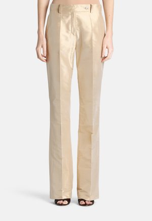 Beige satijnen hoge taille broek met plooien aan de voorkant, knoop- en ritssluiting, gedragen met zwarte open-toe hakken.