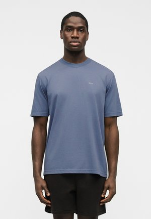 Νεαρός άνδρας που φοράει απλό μπλε κοντομάνικο μπλουζάκι με μικρό λευκό κείμενο στο στήθος και μαύρο σορτς, στέκεται μπροστά σε λευκό απλό φόντο.