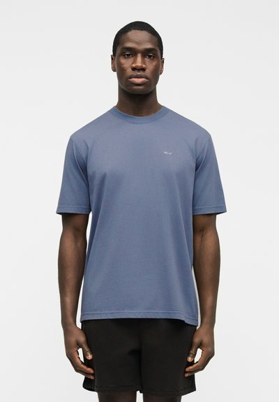 Jeune homme portant un t-shirt bleu uni à manches courtes avec un petit texte blanc sur la poitrine et un short noir, debout devant un fond blanc uni.