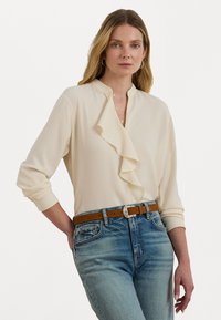 Cream blouse with a ruffled placket and long sleeves, paired with light blue denim jeans and a brown belt with a silver buckle.
