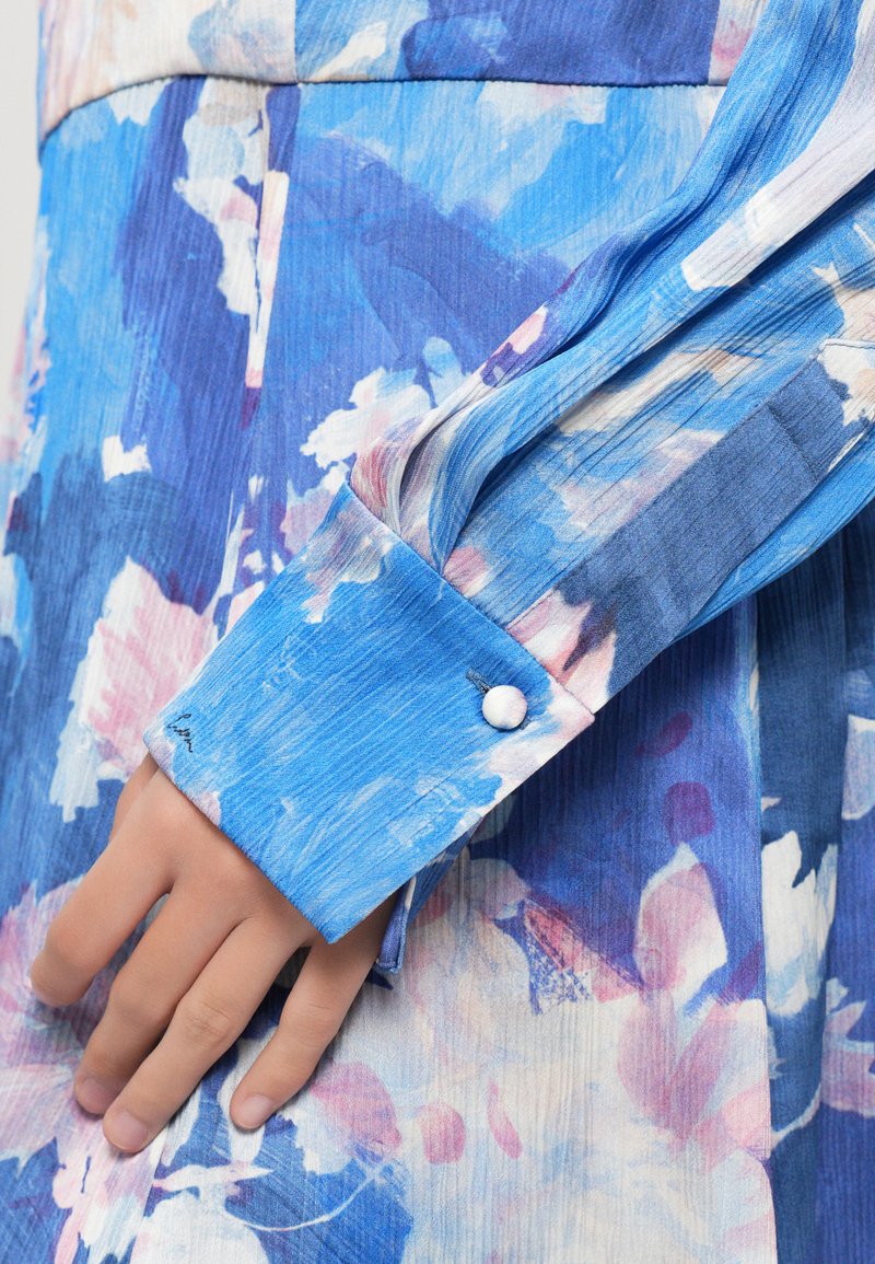 Hand rustend naast de manchetknoop van een blauwe bloemrijke blousejurk met een enkele bedekte knoopsluiting.