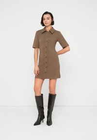 Πλεκτό καφέ φόρεμα με γιακά και κοντά μανίκια, με κουμπιά μπροστά. Φορεμένο με ψηλές μαύρες μπότες. Απλό φόντο.