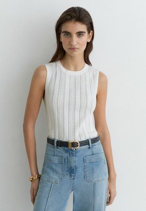Junge Frau mit langen braunen Haaren, die einen ärmellosen weißen strukturierten Top, hellblaue Jeans mit hoher Taille, schwarzen Gürtel und goldenen Armreif trägt.
