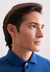 Camicia di cotone blu con colletto e chiusura con bottoni. Primo piano del profilo di una persona, che mette in evidenza la pelle liscia, i capelli scuri e i tratti naturali.