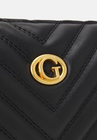 Bolso de mano de cuero negro con costuras en forma de cheurón, adornado con un destacado logo circular dorado. Textura suave con detalles refinados.