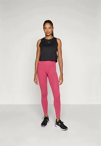 Schwarzes ärmelloses Oberteil und enganliegende rosa Leggings, die den glatten Stoff zur Geltung bringen. Schwarze Laufschuhe mit rosa Akzenten. Das Modell steht frontal.