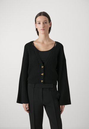 Schwarze gerippte Strickjacke mit drei goldenen Dreiecksknöpfen, weiten Ärmeln und einem tiefen V-Ausschnitt, kombiniert mit einem schwarzen Top und maßgeschneiderten schwarzen Hosen.