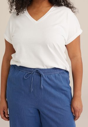 Kobieta ubrana w biały top z krótkim rękawem i dekoltem w serek oraz niebieskie spodnie z elastycznym pasem i sznurkiem, stojąca na neutralnym tle.