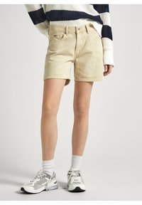 Bermudas de algodón beige con un corte recto, cinco bolsillos y una cintura con un pasador para cinturón. Llevadas con zapatillas y calcetines blancos.