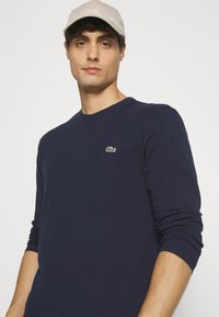 T-shirt à manches longues de couleur marine en coton, avec un col rond et un petit logo Lacoste vert sur la poitrine. Le modèle porte une casquette beige.