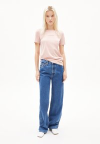 Lichtroze katoenen T-shirt met een subtiele logo, gecombineerd met een wijde blauwe denim jeans. Model draagt witte schoenen en staat tegen een effen achtergrond.