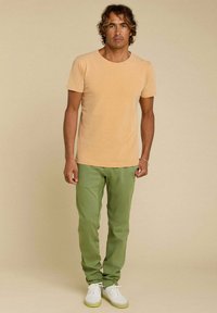 T-shirt à manches courtes, couleur orange clair, associé à un pantalon slim fit vert kaki et des baskets blanches, se tenant sur un fond beige uni.