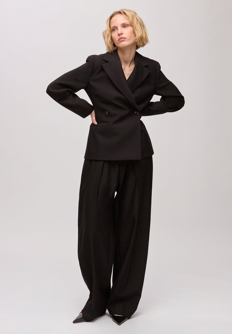 Femme aux cheveux blonds courts portant un blazer noir croisé et un pantalon à jambes larges, debout les mains sur les hanches.